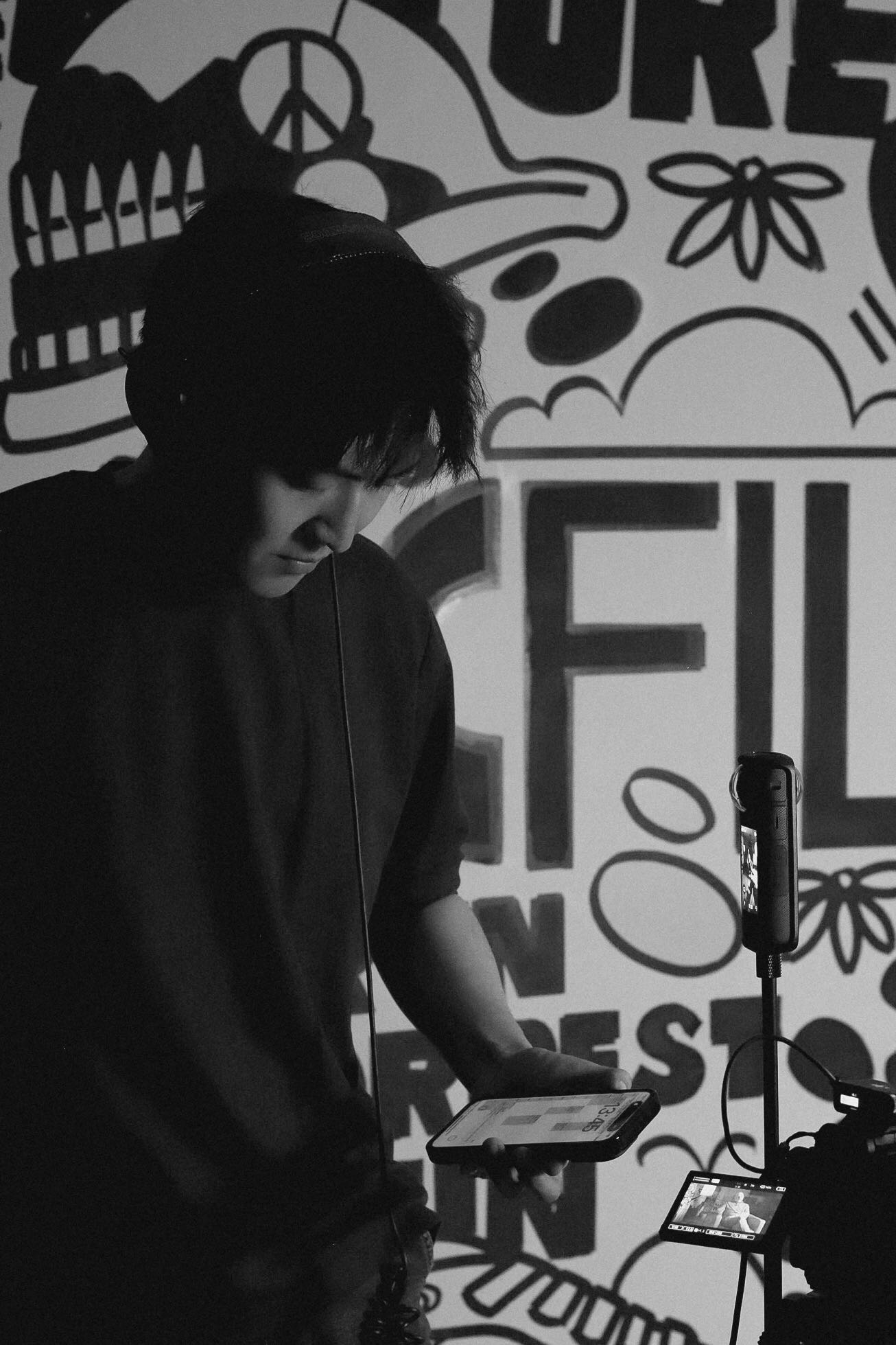 Black-and-white on-set portrait of Boyi Tan checking a phone beside camera gear